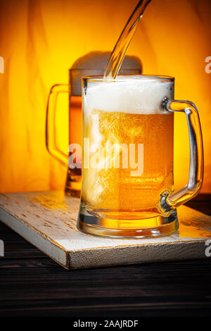 Versare la birra chiara in tazze. Birra Leggera o Birra Light Glass tazze con schiuma bianca sulla tabella Woowen e sfondo giallo Foto Stock