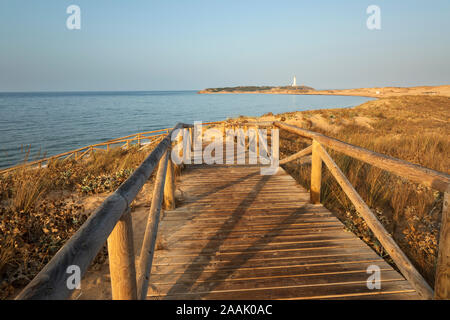 Passerella in legno giù alla spiaggia di Capo Trafalgar con il faro dietro, a Caños de Meca, Costa de la Luz, la provincia di Cadiz Cadice, Andalusia, Spagna Foto Stock