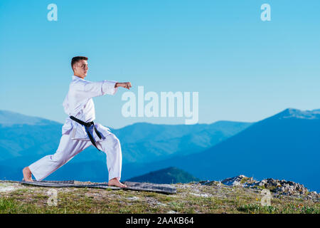 Il Karate uomo in un kimono esegue una mano anteriore kick (Choku-zuki) mentre si sta in piedi sul prato verde sulla cima di una montagna. Foto Stock