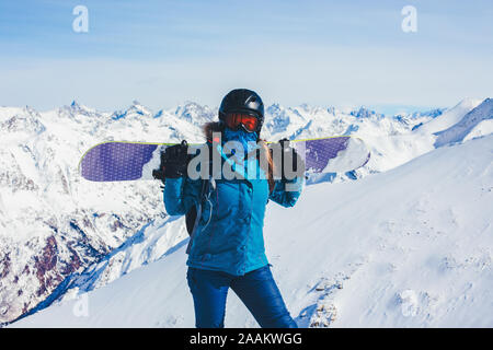 Sfondo sport snowboarder ragazza nel casco e maschera da alte montagne, detiene una snowboard sulle sue spalle. Stile di vita invernali e una estrema Foto Stock