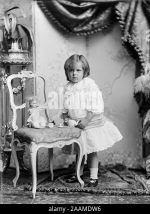 Alate vittoriano o inizio Edwardian inglese vintage fotografia in bianco e nero che mostrano molto una giovane ragazza in un fotografi studios, vestito nella sua migliori vestiti di bianco, in piedi da una sedia che ha sua BAMBOLA su di esso, con Toy tazze e piattini. Foto Stock