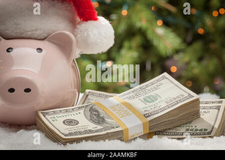 Salvadanaio indossando Santa Hat vicino a pile di centinaia di fatture del dollaro sui fiocchi di neve. Foto Stock