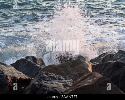 Le onde battendo contro le rocce su una spiaggia rocciosa, spruzzi d'acqua, gocce in un rosa tramonto. Foto Stock