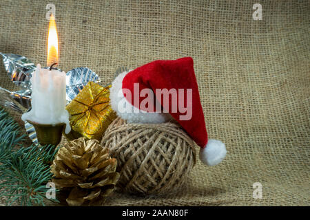 Sfondo di natale con candela che brucia, decorazioni, dono, rotolo di spago e Santa hat su tela rustico sfondo con copia spazio per una vacanza gre Foto Stock