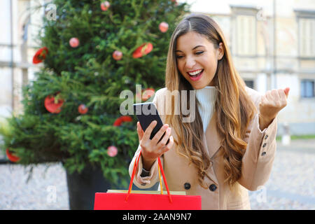 Ragazza di Natale. Felice eccitato giovane donna con le borse della spesa in mano christams di acquisto si presenta con il suo smart phone all'esterno. Foto Stock