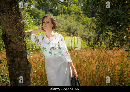 Donna in 40's vestiti sotto un albero di olivo nella campagna abruzzese con un campo di grano in background Foto Stock