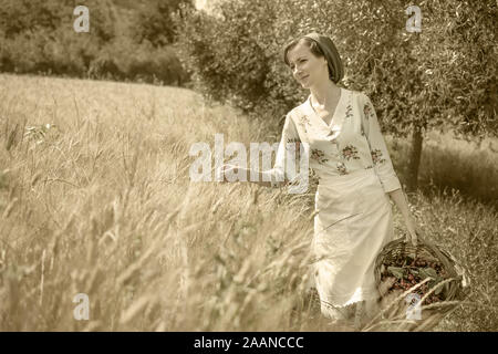 Donna in 40s vestiti passeggiate in campagna italiana, accanto ad un campo di grano, portando un cesto di ciliegie Foto Stock