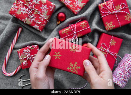 Sfondo di natale con confezioni regalo, bugne di corda e decorazioni su rosso. Preparazione per le vacanze. Vista dall'alto. Donna di mani confezione regalo. Foto Stock