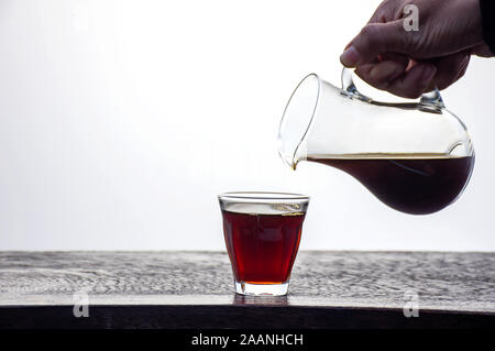 Mano che tiene una brocca di caffè nero versata in un bicchiere su un tavolo di legno. Foto Stock