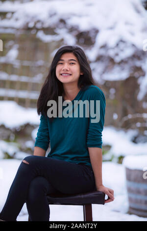 Biracial teen ragazza rilassante su uno sgabello sedile al di fuori in inverno con neve coperto cantiere in background Foto Stock