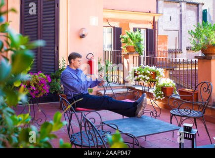 Bel uomo caucasico negli anni quaranta di relax all'aperto sul rosso terracotta veranda italiana con le gambe sul tavolo, guardando a smartphone Foto Stock