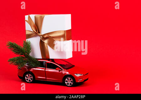 Sfondo di natale. Rosso auto giocattolo con albero di Natale ramo di abete e la presente confezione regalo su sfondo rosso Foto Stock