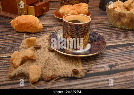 Una tazza di caffè, pezzi di zucchero di canna in una zuccheriera e croissant su uno sfondo di legno Foto Stock