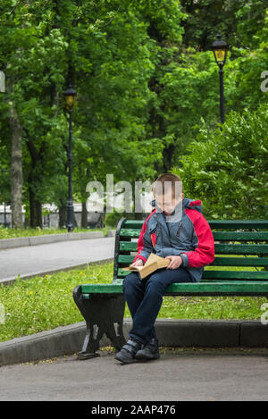 Ragazzo sorridente in un abbigliamento informale è la lettura di un libro. Il ragazzo è seduto da solo su una vecchia panca di legno nel parco. Messa a fuoco selettiva. Non focalizzato parco verde a backgroun Foto Stock