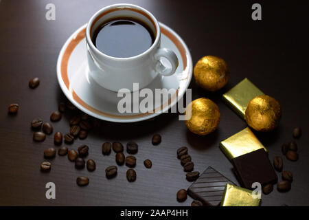 Tazza di caffè con fagioli sparsi intorno e barrette di cioccolato, dolci e dolciumi in involucri di oro su legno bruno business desk, close up, copia di spazio, Foto Stock