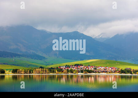 Villaggio sulle rive di un lago. bellissimo paesaggio rurale in montagna. lontane cime coperte di neve in nubi. fantastico scenario della western Alti Tatra i Foto Stock