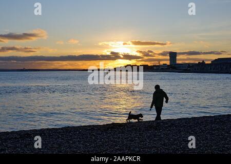 Silhouette di un uomo a piedi un cane sulla spiaggia a Portsmouth contro un tramonto in inverno, Hampshire England Regno Unito Foto Stock