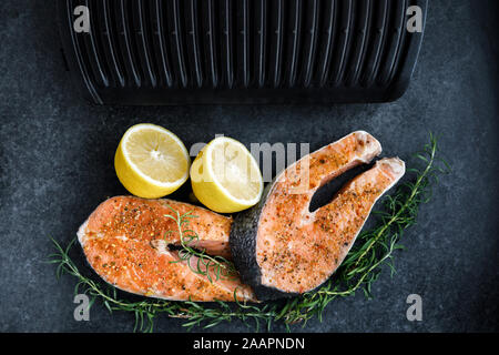 Preparazione per la frittura un salmone fresco bistecca su una griglia elettrica contro uno sfondo scuro, lungo con limone e rosmarino. Foto Stock