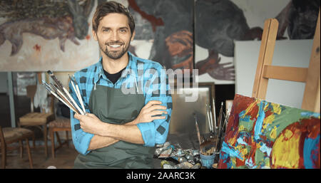 Ritratto di giovane artista sorridente in posa con dei pennelli nel suo loft studio Foto Stock