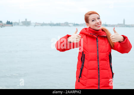 Donna all'aperto che mostra come il pollice in alto gesto con entrambe le mani. Giovani redhead lady indossa cappotto rosso in piedi fuori, all'aperto in autunno e inverno. Città Foto Stock