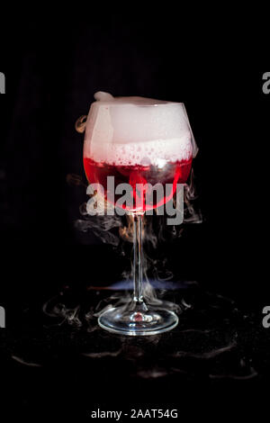 Un bicchiere di rosso cocktail con ghiaccio secco fumo e sfondo nero Foto Stock