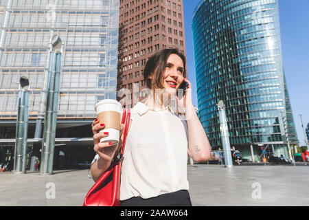 Sorridente giovane imprenditrice con caffè da asporto al telefono nella città di Berlino, Germania Foto Stock