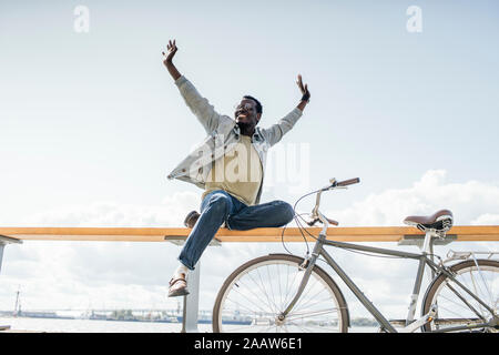 Giovane uomo con la bicicletta, seduti su railng dal mare, fingendo di volare Foto Stock