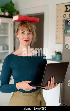 Ritratto di donna sorridente in piedi con il computer portatile in cucina il bilanciamento libro sulla sua testa Foto Stock
