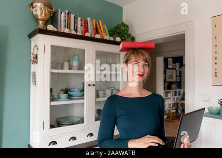 Ritratto di donna sorridente in piedi con il computer portatile in cucina il bilanciamento libro sulla sua testa Foto Stock