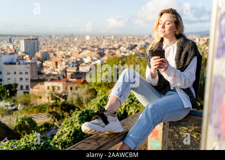 Giovane donna seduta sulla ringhiera sopra l'azienda città telefono cellulare, Barcellona, Spagna Foto Stock