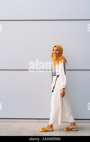 Giovane donna musulmana che indossa il hijab giallo e a piedi nella parte anteriore di un muro bianco Foto Stock