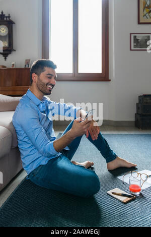 Ritratto di giovane sorridente uomo seduto a piedi nudi sul pavimento a casa guardando smartphone Foto Stock