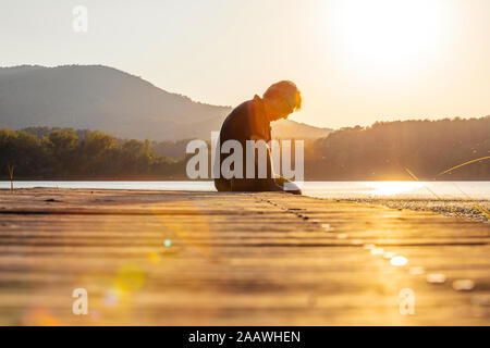 Senior uomo seduto su una passerella in legno e guardando verso il basso contro il sole Foto Stock