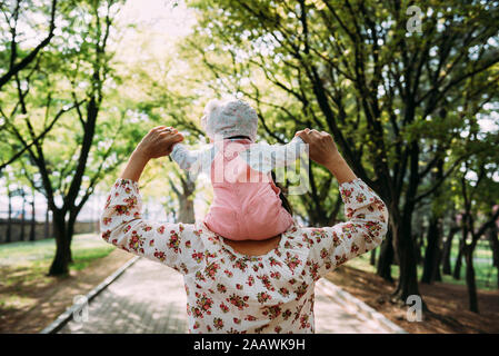 Madre camminando in un parco con la bambina sulle sue spalle Foto Stock