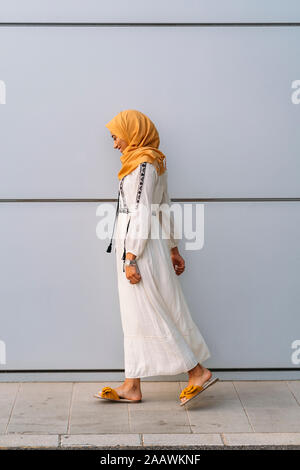 Giovane donna musulmana che indossa il hijab giallo e a piedi nella parte anteriore di un muro bianco Foto Stock