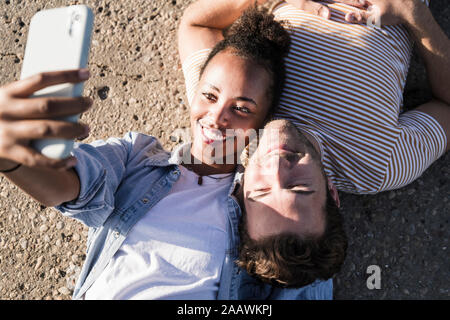 Felice coppia giovane sdraiato sul pavimento di cemento prendendo un selfie Foto Stock