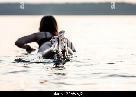 Vista posteriore della giovane donna sdraiata sulla tavola da surf da Sunset, il lago di Starnberg, Germania Foto Stock