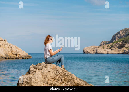 Redheaded donna seduta su roccia e la rivista di lettura Foto Stock