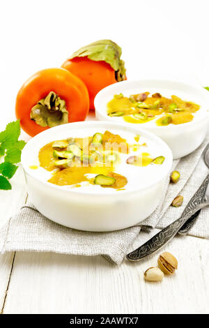 Il dessert di yogurt, persimmon e miele con vaniglia, il cardamomo e i pistacchi in due ciotole su un asciugamano, menta e cucchiaio sullo sfondo della luce boa in legno Foto Stock
