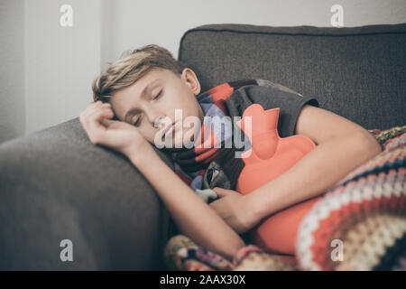 Ragazzo malato a letto con coperta di lana e bottiglie di acqua calda. Triste teen con l'influenza si appoggia a casa da solo in una fredda giornata invernale. Bambino con infettare stagionali Foto Stock