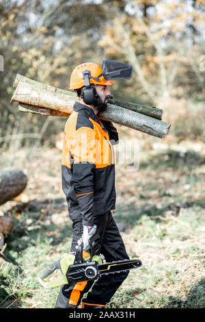 Professional lumberman in indumenti da lavoro protettiva con una motosega che trasportano tronchi di legno durante il lavoro sul disboscamento in pineta Foto Stock