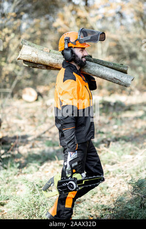 Professional lumberman in indumenti da lavoro protettiva con una motosega che trasportano tronchi di legno durante il lavoro sul disboscamento in pineta Foto Stock