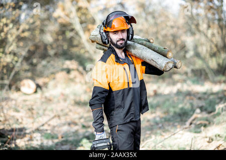 Ritratto di un professionista lumberman in indumenti da lavoro protettiva con un chainsaw e tronchi di legno durante il lavoro sul disboscamento in pineta Foto Stock