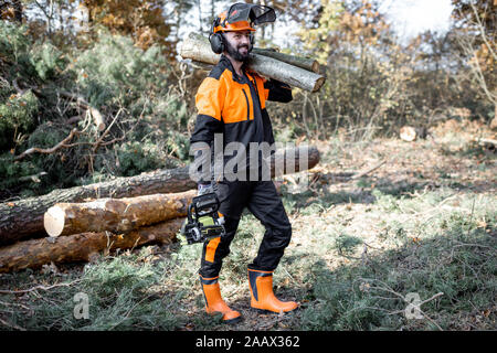 Professional lumberman in indumenti da lavoro protettiva con una motosega che trasportano tronchi di legno durante il lavoro sul disboscamento in pineta Foto Stock