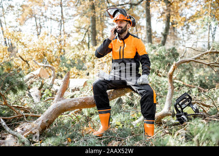 Professional lumberman in indumenti da lavoro protettiva parlando al telefono mentre è seduto sulla struttura ad albero abbattuto, riposo dopo il duro lavoro della foresta Foto Stock