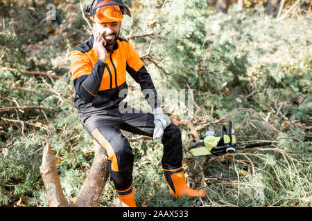 Professional lumberman in indumenti da lavoro protettiva parlando al telefono mentre è seduto sulla struttura ad albero abbattuto, riposo dopo il duro lavoro della foresta Foto Stock