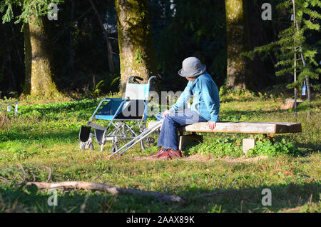 Una donna anziana con una sedia a rotelle seduta su una panchina nel parco. Foto Stock