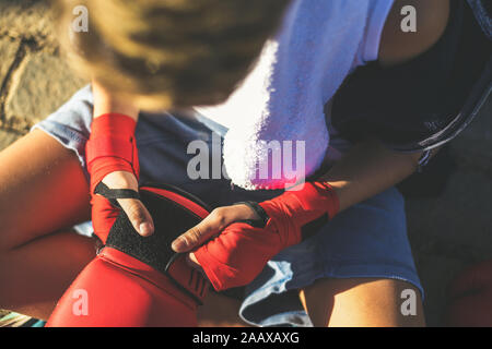 Bel giovane maschio con bande di inscatolamento nelle sue mani, guardando i guanti seduti sulla strada. Il ragazzo si prepara per un allenamento boxe, ascoltando musica. Sport, gioventù Foto Stock