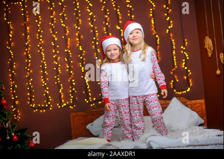 Due graziosi Piccole sorelle di santa dei tappi sono abbracciando mentre in una stanza su un letto contro uno sfondo di brillanti luci festose. Attesa per la hol Foto Stock