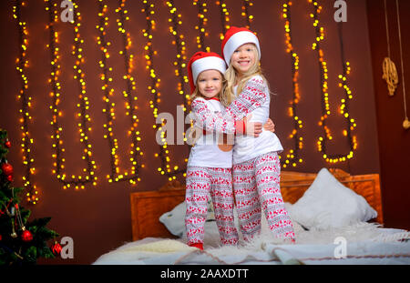 Due graziosi Piccole sorelle di santa dei tappi sono abbracciando mentre in una stanza su un letto contro uno sfondo di brillanti luci festose. Attesa per la hol Foto Stock